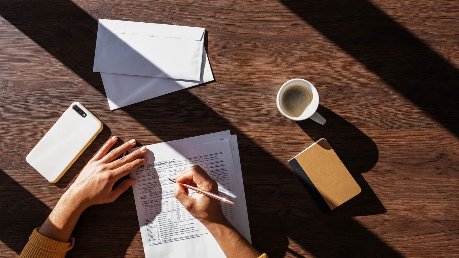 Woman writing on a piece of paper on a desk with a coffee cup, a smartphone and som envelopes on it